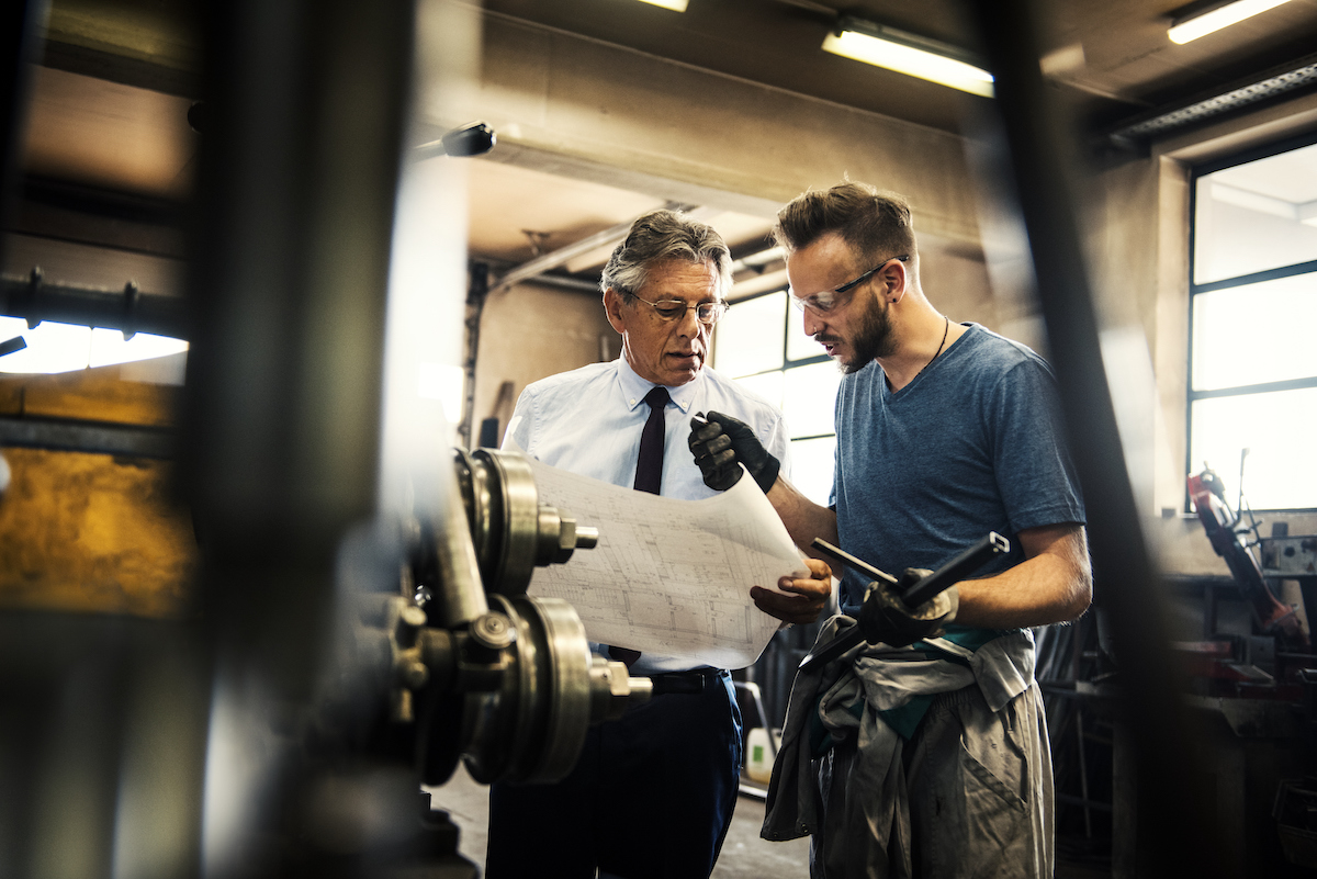 Businessman and welder examining blueprints on workshop floor. 